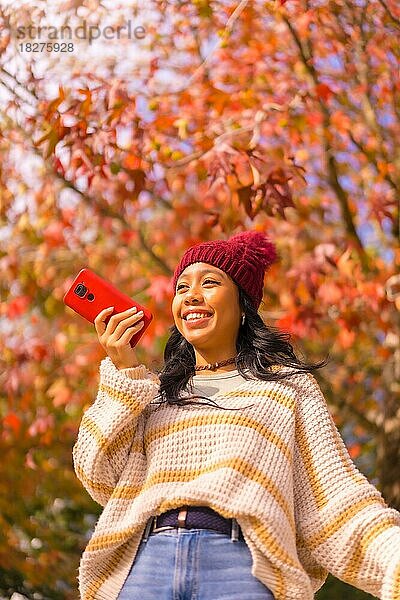 Asiatisches Mädchen im Herbst mit einem Handy  das lächelnd eine Sprachnachricht in einem Wald aus roten Blättern sendet  Technik