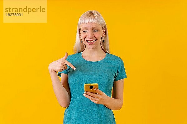 Lächelnd auf das Telefon zeigend  blondes kaukasisches Mädchen im Studio auf gelbem Hintergrund