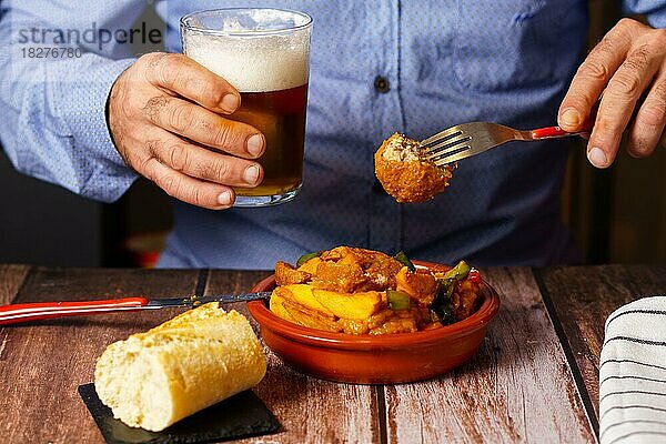 Mann mit Bart und blauem Hemd isst Fleischbällchen mit Spaghetti in einem Restaurant mit einem Glas Bier in der Hand