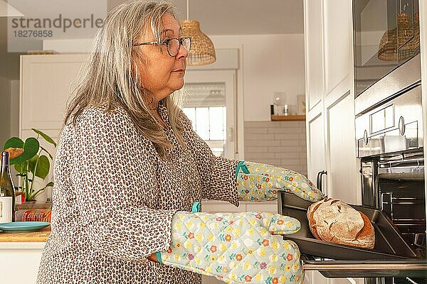 Ältere weißhaarige Frau  die in ihrer Küche zu Hause Brot in den Ofen schiebt