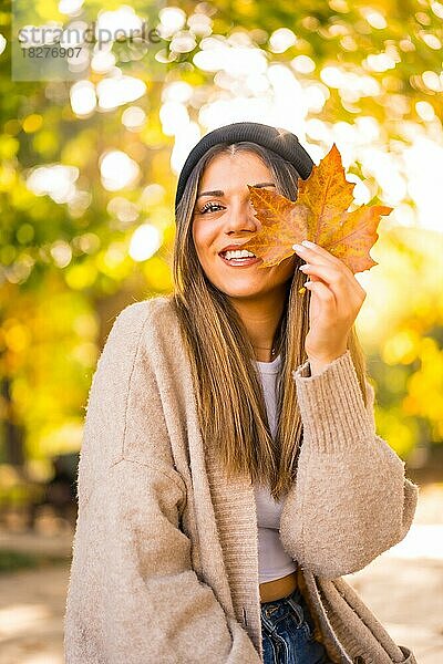 Junges blondes Mädchen mit Wollmütze im Herbst  mit einem Blatt auf dem Gesicht in den Sonnenuntergang lächelnd