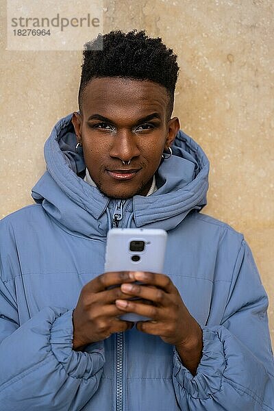 Porträt eines schwarzen ethnischen Mannes mit einem Telefon und einer blauen Jacke auf einem gelben Hintergrund  der auf soziale Netzwerke oder die digitale Presse schaut