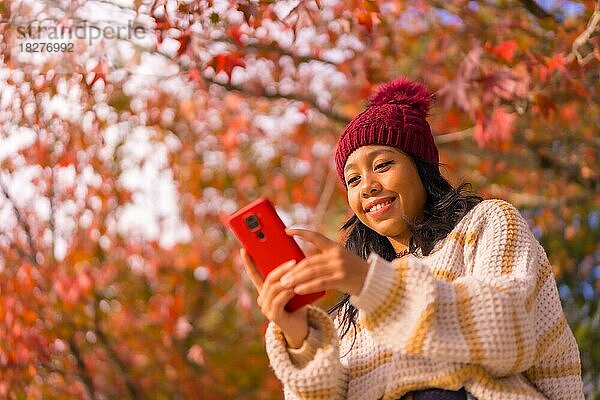 Asiatisches Mädchen im Herbst mit einem Handy  das eine Nachricht in einem Wald aus roten Blättern schreibt  Technik