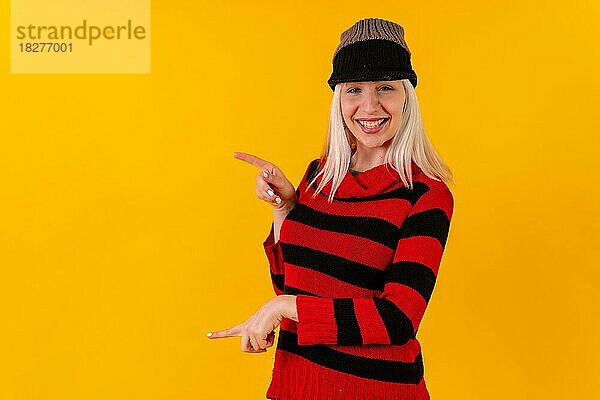 Nach links weisendes blondes kaukasisches Mädchen mit Wollmütze auf gelbem Hintergrund Studio