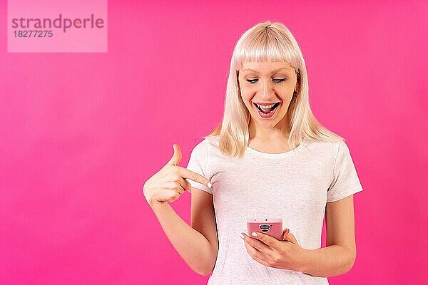 Blondes kaukasisches Mädchen im Studio auf rosa Hintergrund  Kopierraum  lächelnd auf das Handy zeigend