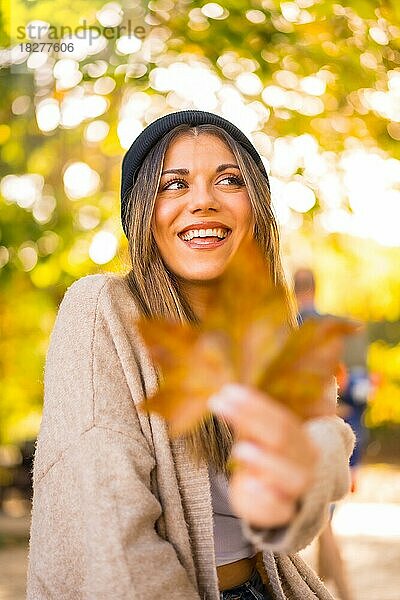 Junges blondes Mädchen mit Wollmütze im Herbst  mit einem Blatt auf dem Gesicht in den Sonnenuntergang lächelnd