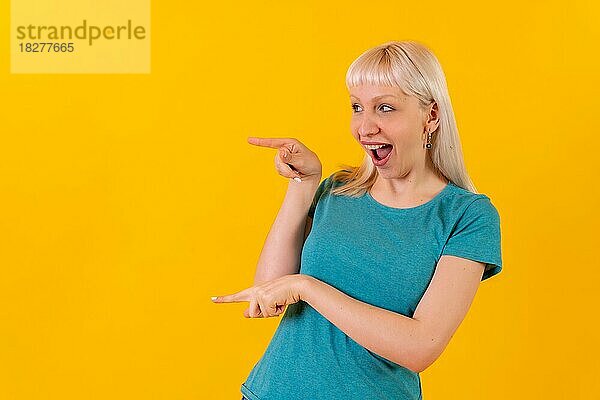 Portrait lächelnd nach links zeigend  blondes kaukasisches Mädchen im Studio auf gelbem Hintergrund