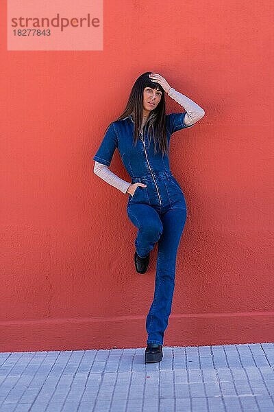 Posieren eines lächelnden brünetten Mädchens  das in einem blauen Jeans-Outfit an einer Wand lehnt. Hübsche Kaukasierin posiert vor einem roten Hintergrund