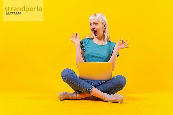 Lächelnd sitzen mit einem Laptop tun ok  blonde kaukasische Mädchen im Studio auf gelbem Hintergrund