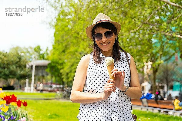 Eine touristische Frau  die die Stadt besucht  isst eine Eistüte  lächelt und genießt den Urlaub  Solo weibliche Reisende Konzept  trägt eine Sonnenbrille und einen Hut