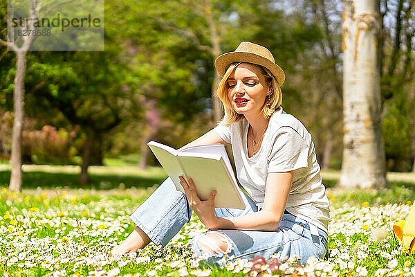 Ein junges blondes Mädchen mit Hut liest im Frühling in einem Park in der Stadt ein Buch  macht Urlaub in der Natur und neben Gänseblümchen  sitzt im Gras