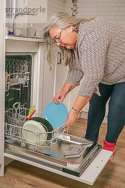Ältere weißhaarige Frau  die in ihrer Küche zu Hause Geschirr in den Geschirrspüler einräumt
