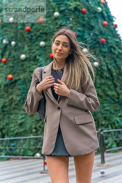 Porträt am Weihnachtsbaum in der Stadt  lächelnde junge Frau im Trenchcoat