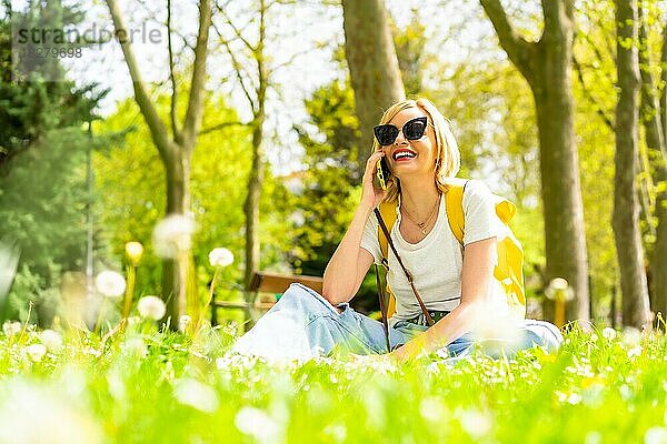 Eine lächelnde blonde Touristin  die ihre Freunde mit einem Telefon anruft  trägt einen Hut und eine Sonnenbrille und sitzt im Frühling im Gras neben Gänseblümchen in einem Park in der Stadt