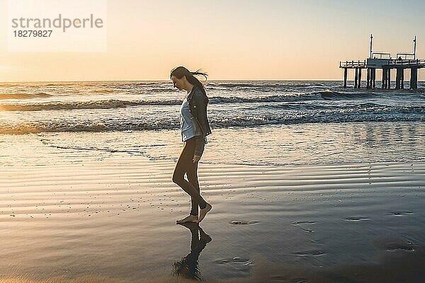 Eine einsame Frau am Strand  die bei Sonnenaufgang barfuß am Ufer entlang läuft. Raum kopieren