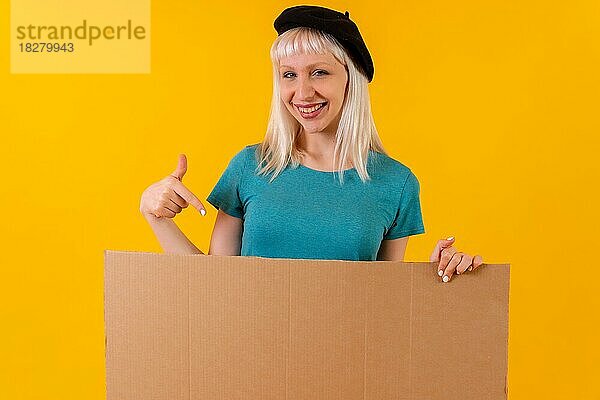 Lächelnd und auf ein leeres Werbeplakat zeigend  blondes kaukasisches Mädchen im Studio auf gelbem Hintergrund