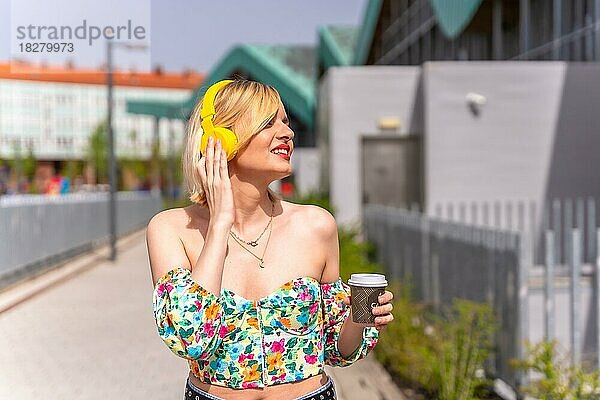Eine hübsche blonde Frau  die mit gelben Kopfhörern und einem Kaffee zum Mitnehmen in der Stadt Musik hört  Lebensstil eines Models in der Stadt