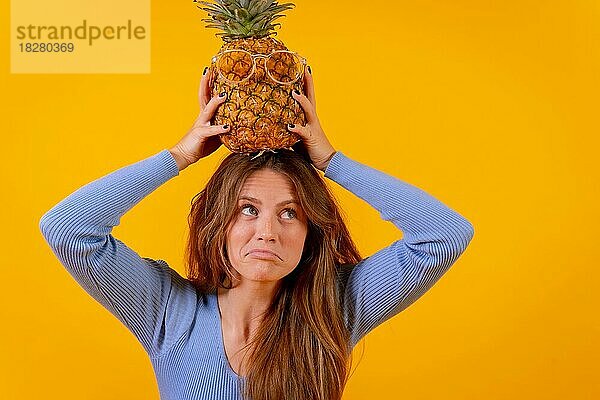 Frau mit Ananas und Sonnenbrille in einem Studio auf gelbem Hintergrund  mit Blick nach oben