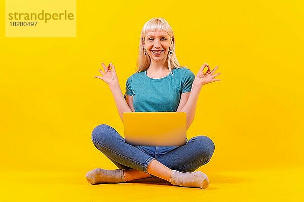 Lächelnd sitzend mit einem Laptop  blonde kaukasische Mädchen im Studio auf gelbem Hintergrund