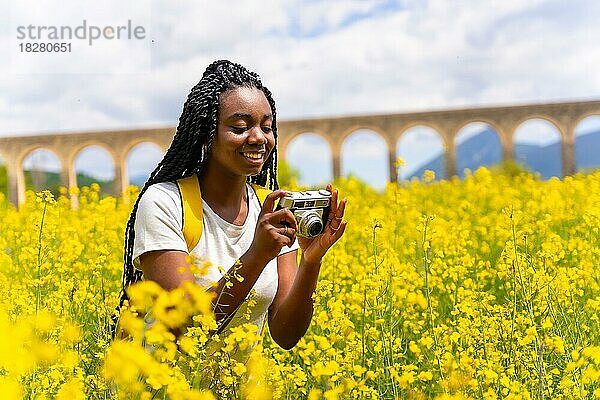 Fotografieren von Blumen mit einer alten Kamera  ein schwarzes ethnisches Mädchen mit Zöpfen  eine Reisende  in einem Feld mit gelben Blumen