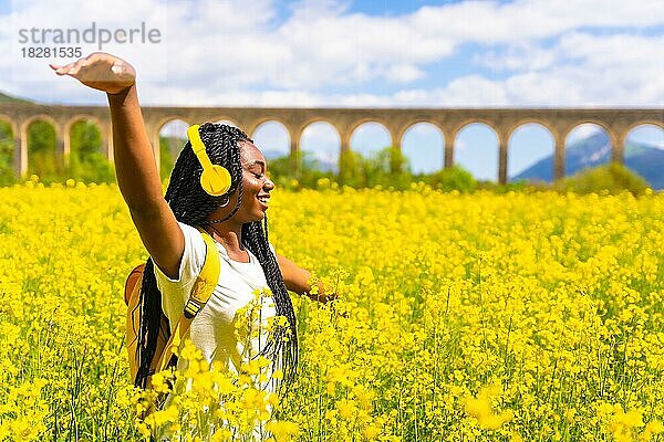 Ein Mädchen schwarzer Herkunft mit Zöpfen  eine Reisende  tanzt zu Musik mit gelben Kopfhörern und geschlossenen Augen in einem gelben Blumenfeld