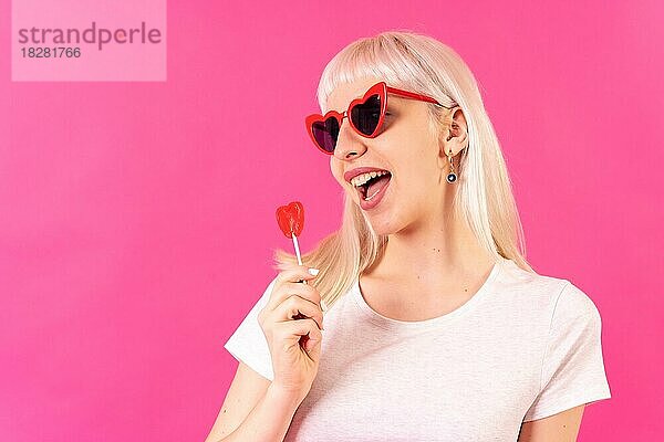 Blonde kaukasische Mädchen im Studio auf rosa Hintergrund  lächelnd in Gläser von Herzen mit einem Lutscher
