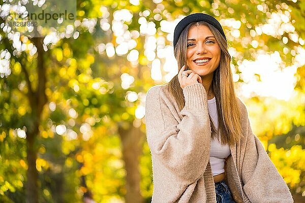 Ein junges blondes Mädchen mit einer Wollmütze im Herbst  das bei Sonnenuntergang lächelnd im Park sitzt