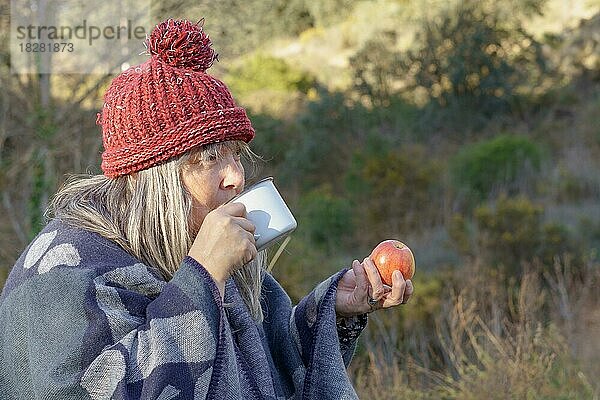 Frau mit weißen Haaren und roter Mütze trinkt Kaffee und isst einen Apfel auf einem Feld mit Bäumen im Hintergrund