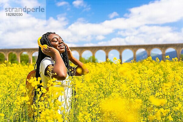 Musik hörend mit gelben Kopfhörern  ein schwarzes ethnisches Mädchen mit Zöpfen  eine Reisende  in einem Feld mit gelben Blumen