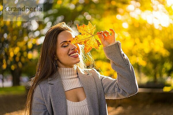 Eine hübsche Frau genießt den Herbst in einem Park bei Sonnenuntergang  mit einem Blatt auf ihrem Gesicht