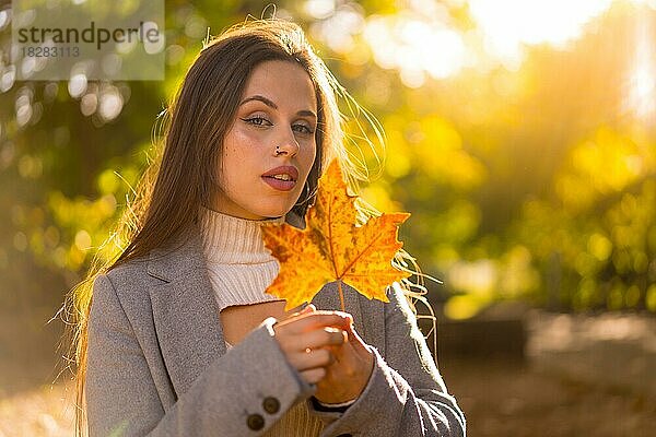 Eine hübsche Frau genießt den Herbst in einem Park bei Sonnenuntergang  mit einem Blatt vom Baum