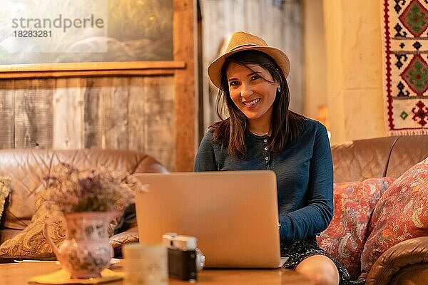 Eine hübsche Touristin mit Hut und Fotoapparat  die in einer Cafeteria Tee trinkt und im Urlaub mit einem Computer aus der Ferne arbeitet