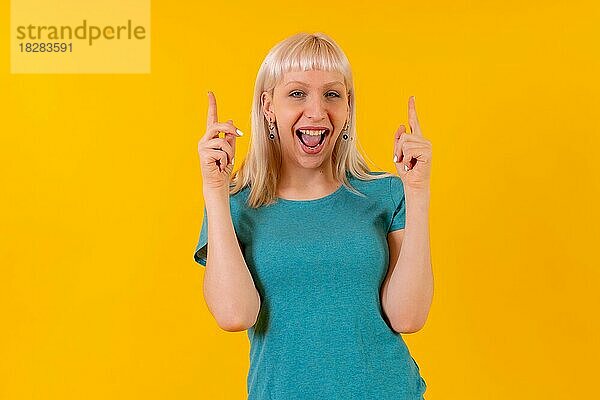 Porträt lächelnd nach oben zeigend  blondes kaukasisches Mädchen im Studio auf gelbem Hintergrund