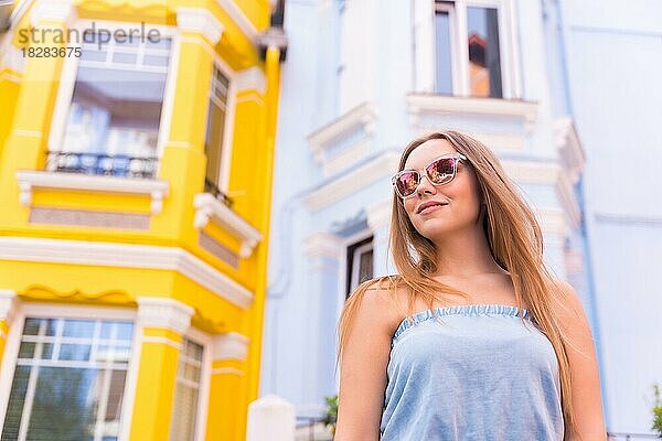 Porträt einer attraktiven jungen Frau mit Sonnenbrille  hinter gelber und blauer bunter Fassade