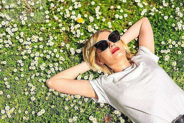 Ein junges blondes Mädchen mit Hut und Sonnenbrille atmet im Frühling in einem Park in der Stadt  Natur  auf dem Gras liegend neben Gänseblümchen  Teil Kopie Raum