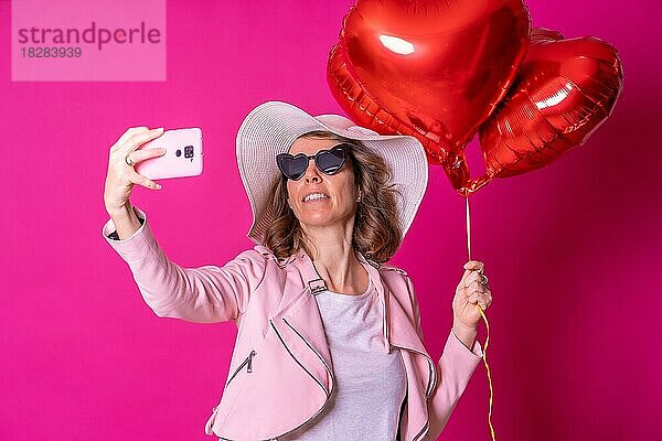 Eine blonde kaukasische Frau  die sich mit einem weißen Hut und einer Sonnenbrille in einem Nachtclub mit einigen Herzluftballons amüsiert und ein Selfie mit ihrem Mobiltelefon macht  rosa Hintergrund