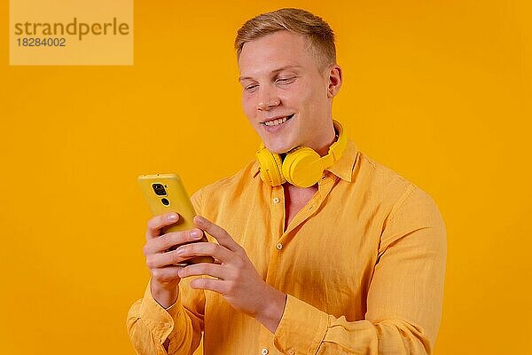 Junger blonder kaukasischer Mann auf gelbem Hintergrund  der die Musik auf dem Handy auswählt