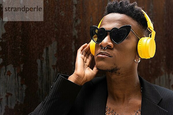 Ein schwarzer ethnischer Mann hört Musik über kabellose gelbe Kopfhörer  trägt eine Herz-Sonnenbrille und hört Musik auf digitalen Plattformen
