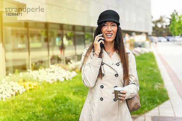 Geschäftsfrau im Trenchcoat mit einem Kaffee in der Hand und einem Gespräch am Telefon