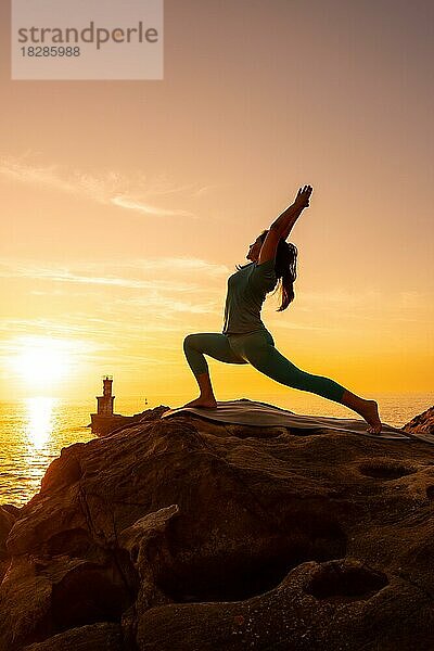 Anjaneyasana  Eine Frau macht Meditations- und Yogaübungen auf einem Felsen bei Sonnenuntergang neben einem Leuchtturm im Meer  gesundes und naturistisches Leben