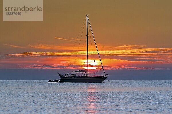 Segelboot  Yacht mit Beiboot vor Anker mit gesenkten Segeln  Silhouette gegen orange Himmel bei Sonnenuntergang