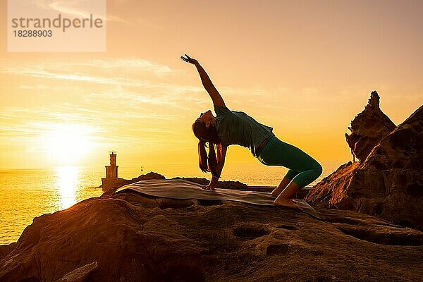 Urdhva Dhanurasana  eine Frau bei Meditations- und Yogaübungen auf einem Felsen bei Sonnenuntergang neben einem Leuchtturm im Meer  gesundes und naturistisches Leben