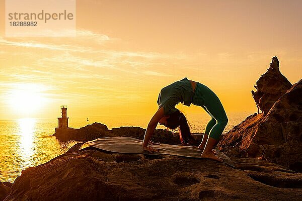 Urdhva Dhanurasana  eine Frau bei Meditations- und Yogaübungen auf einem Felsen bei Sonnenuntergang neben einem Leuchtturm im Meer  gesundes und naturistisches Leben