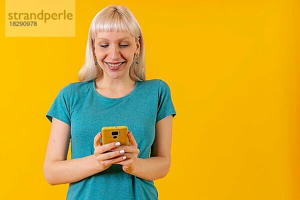 Lächelnd mit Telefon in den Händen  blonde kaukasische Mädchen im Studio auf gelbem Hintergrund