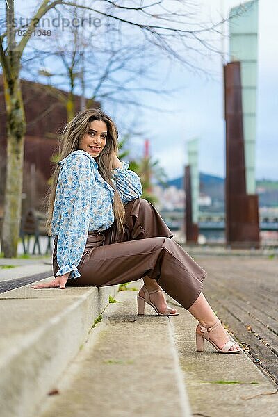 Junge Frau sitzt in einem Park in der Stadt und lächelt  Lifestyle-Konzept  blaues Hemd und braune Hose