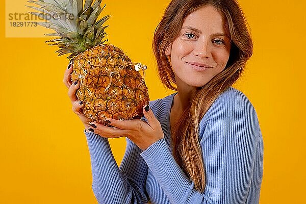 Frau lächelnd mit einer Ananas in Sonnenbrille in einem Studio auf einem gelben Hintergrund  gesund  Sommer