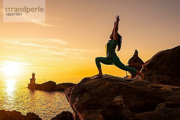 Anjaneyasana  eine Frau bei Meditations- und Yogaübungen auf einem Felsen bei Sonnenuntergang neben einem Leuchtturm im Meer  gesundes und naturistisches Leben