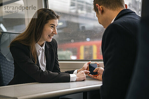 Glückliche Geschäftsfrau zeigt und teilt ihr Smartphone mit einem Kollegen im Zug