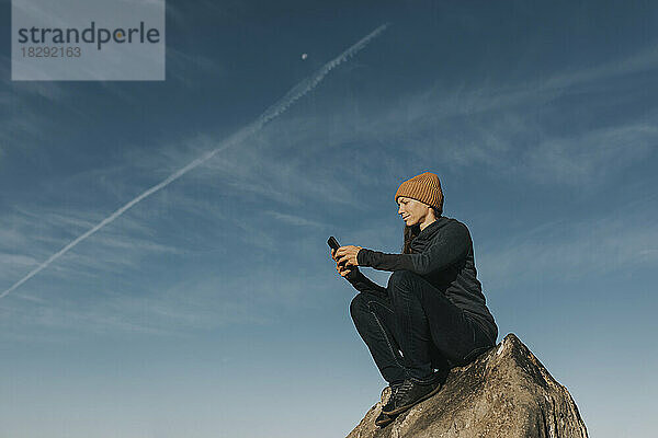 Frau sitzt mit Smartphone auf einem Felsen vor blauem Himmel