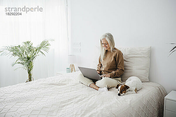 Lächelnde reife Frau benutzt Laptop mit Hund zu Hause im Bett